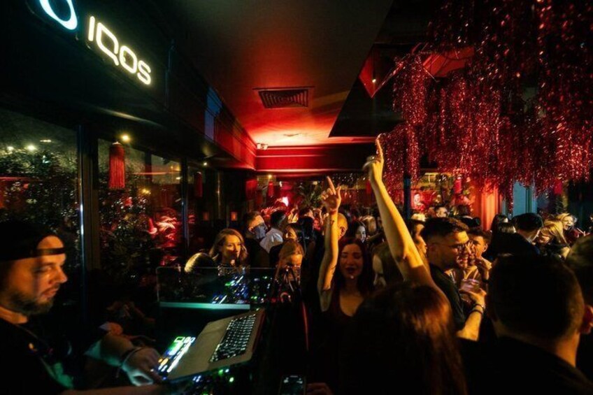 A crowded indoor venue with people dancing and a DJ booth. 1