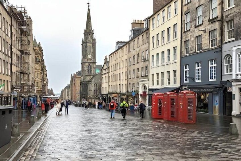A wet cobblestone street lined with historic buildings, a church spire, and red telephone booths. 2