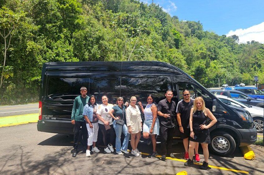 A group of people posing in front of a black van in a parking lot with trees in the background. 1