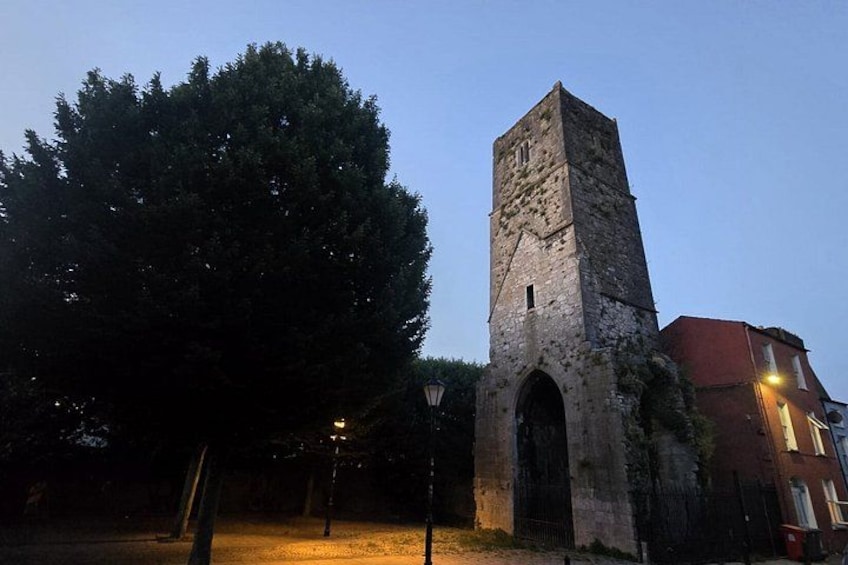 A stone tower with an arched entrance, a large tree, and a building with a red roof in the background. 4