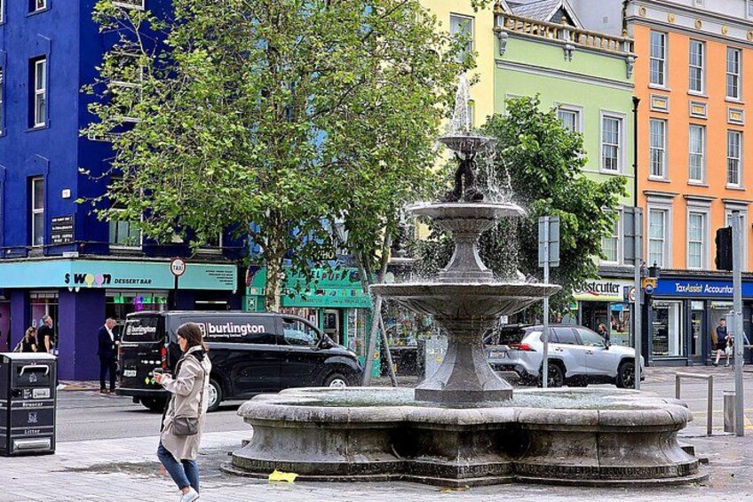 A woman walking past a fountain, with colorful buildings and parked cars in the background. 2
