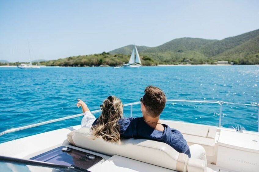 A boat on a clear blue sea with a sailboat in the distance and a hilly landscape in the background. 1