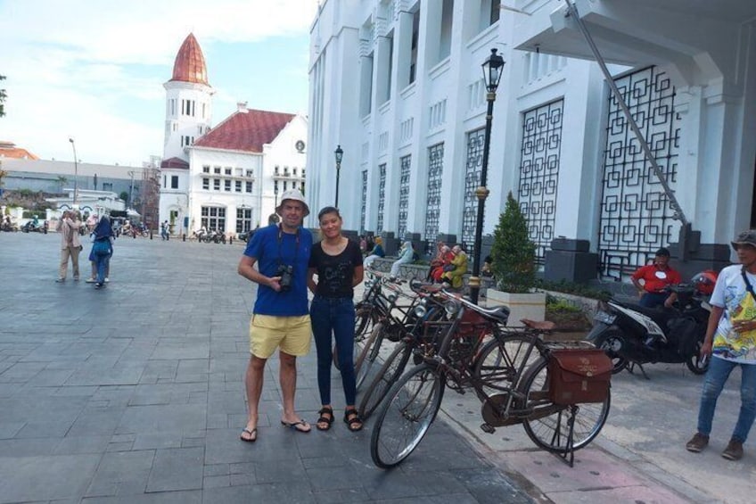 A couple posing in front of a building with a red-tiled roof and a white facade, with bicycles parked nearby. 2
