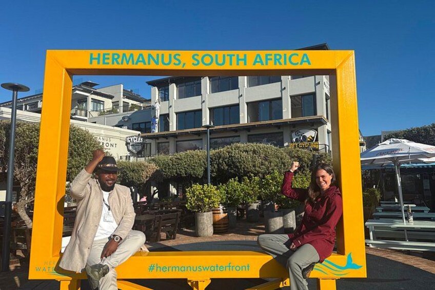 A yellow frame with 'Hermanus, South Africa' written on it, a man and a woman sitting inside, and a hashtag '#hermanuswaterfront'. 3