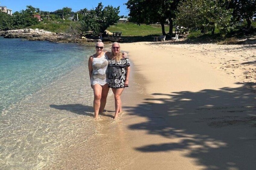 Two people standing at the edge of a beach with clear water and a sandy shore. 2