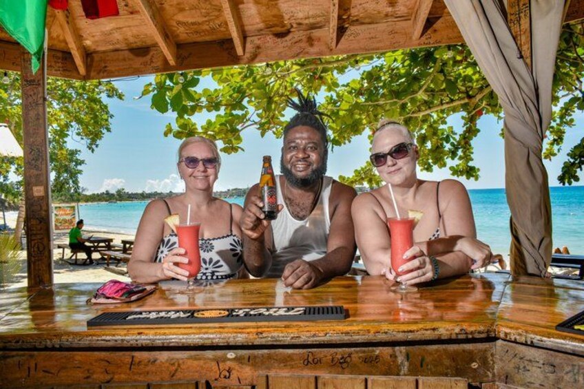 Three people sitting at a wooden table with drinks, a beach and ocean in the background. 3