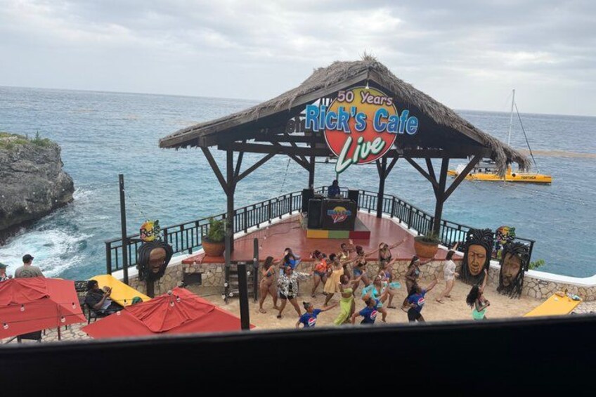 A beachside stage with a 'Rick's Cafe Live' sign, surrounded by people and a boat in the background. 2