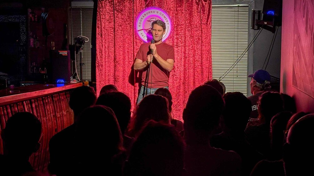 A person performing stand-up comedy on stage with a red curtain backdrop and a neon sign. 1
