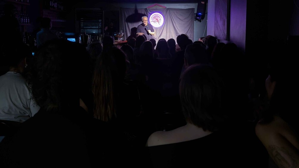 A person speaking to an audience in a dimly lit room with a microphone stand. 2