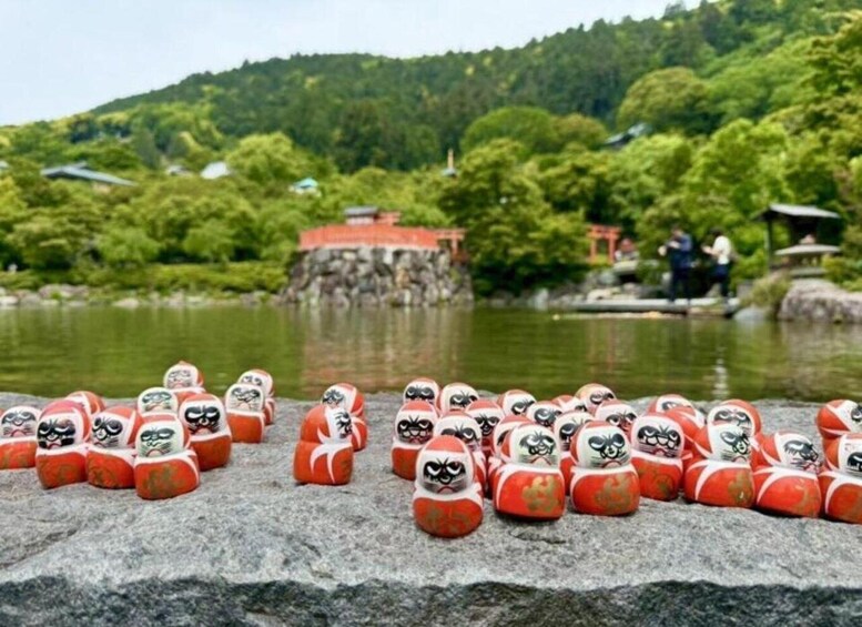 Eine Ansammlung aus roten und weißen Puppen auf einer Steinoberfläche, am Ufer eines Sees mit einem bewaldeten Hügel im Hintergrund. 2