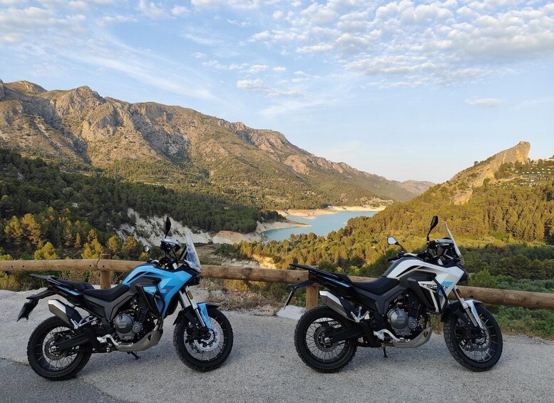 Dos motocicletas estacionadas en un camino de grava, con una vista panorámica de un lago y montañas al fondo. 4