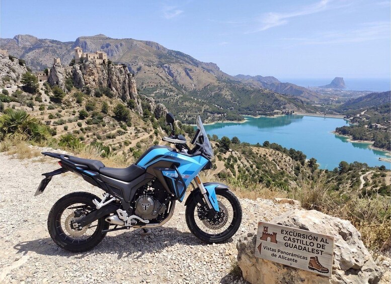 Una motocicleta azul estacionada en un camino de grava con una vista panorámica de un lago, montañas y un castillo al fondo. 1