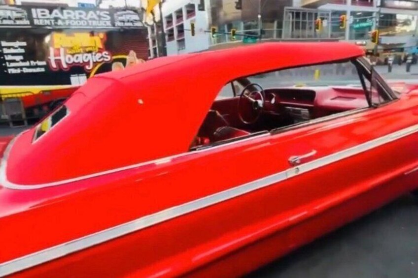 A red vintage car with a white stripe, parked on a street with buildings and a sign for 'Hoagies' in the background. 3