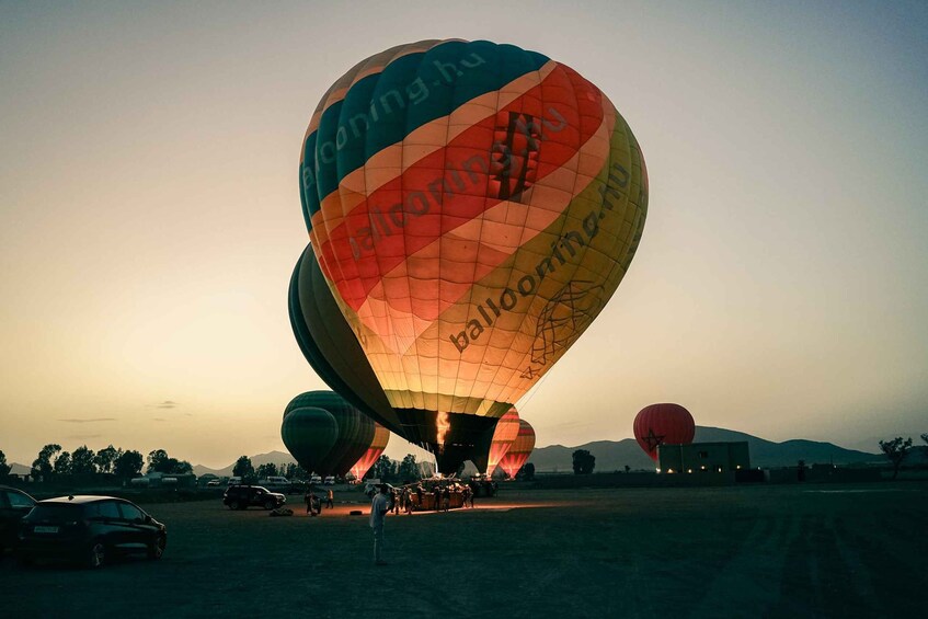 A hot air balloon with multiple colors and text, parked on a field with other balloons and vehicles in the background. 2