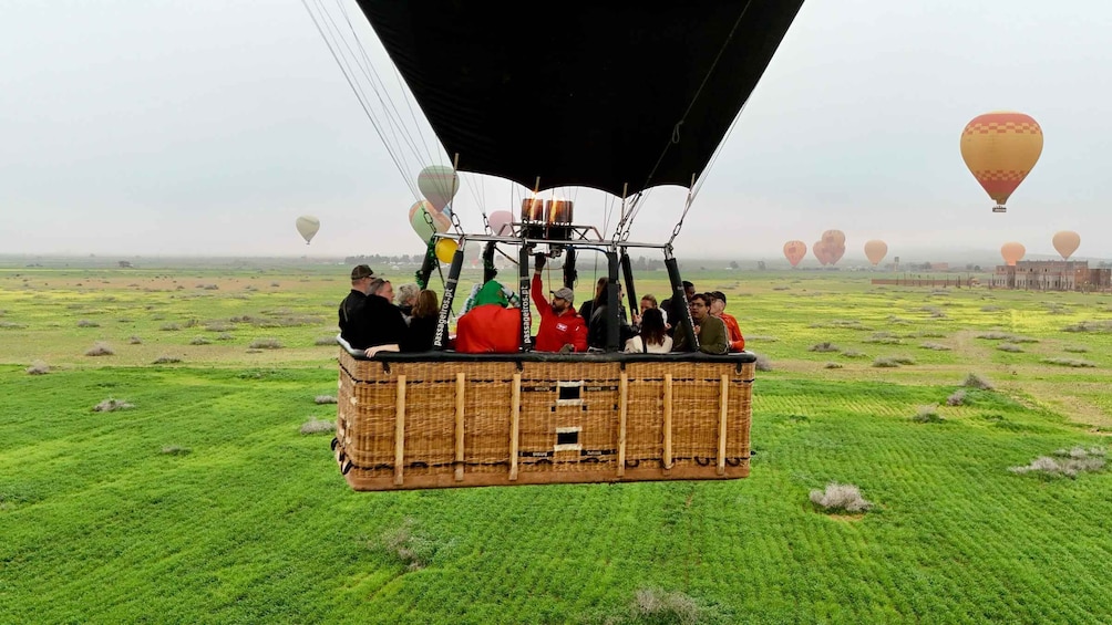 A wicker hot air balloon with passengers, flying over a green field with other balloons in the distance. 1