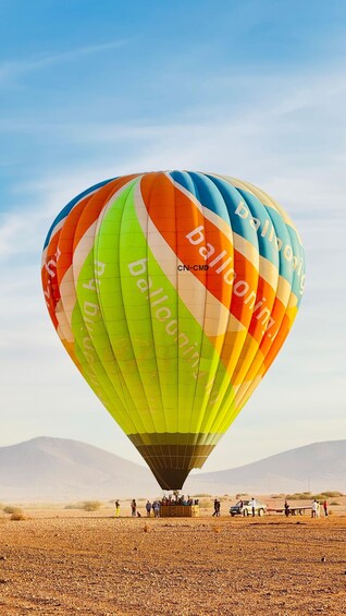 A colorful hot air balloon with the text 'ballooning' and 'CN-CMD' on its side, parked on a dry, rocky ground with a clear sky and mountains in the background. 3