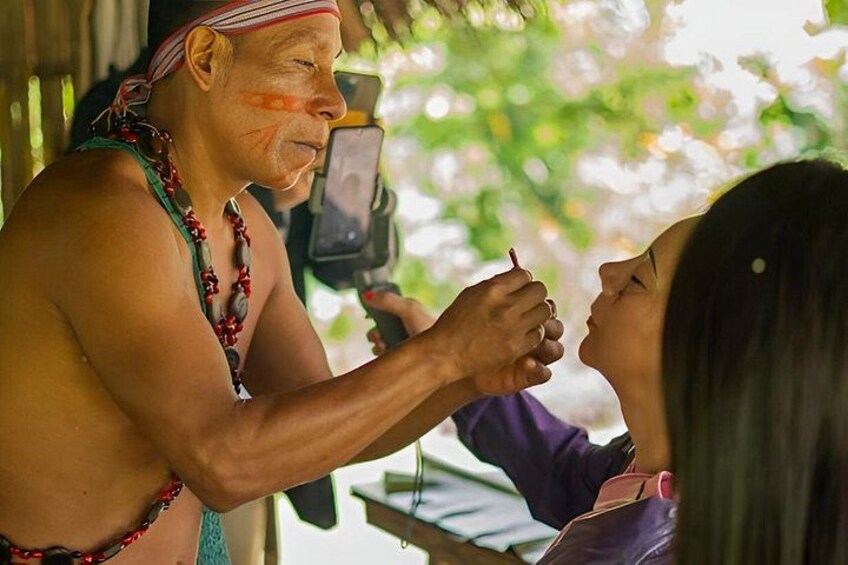 An elderly man and a young woman in a tropical setting, possibly a village, with a camera being used to take a photo. 1