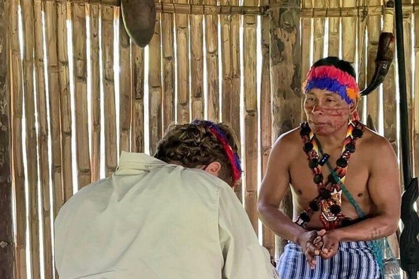 A person in traditional attire with a headdress and beaded necklace, standing in front of a wooden structure. 5