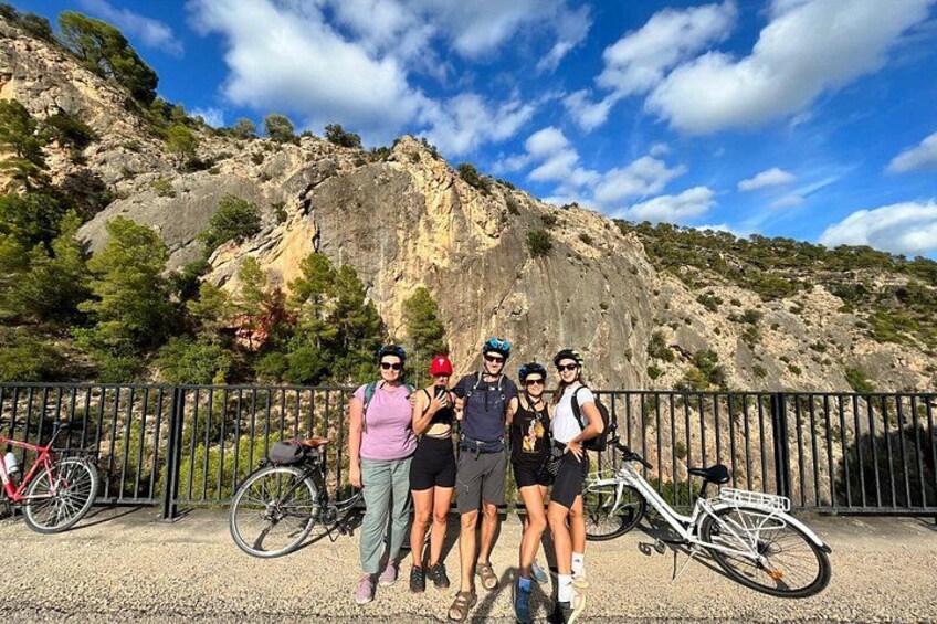 A group of people standing in front of a rocky mountain with bicycles parked behind a metal fence. 5