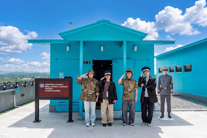 Un grupo de personas con uniforme militar posando frente a un edificio azul con un cartel que indica el Edificio de la Conferencia de la Comisión Militar de Armisticio del Comando de las Naciones Unidas. 1