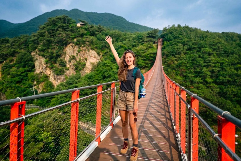 A person on a suspension bridge with red railings, surrounded by lush green mountains. 2