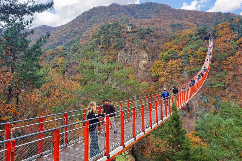 A suspension bridge with red railings, surrounded by autumn foliage and a mountainous backdrop. 3