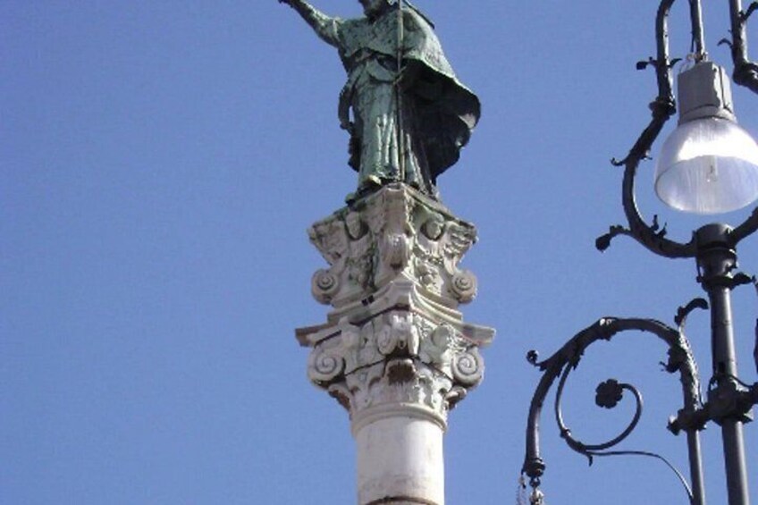 A statue atop a column with intricate carvings, a street lamp with a clear glass shade, and a clear blue sky in the background. 5