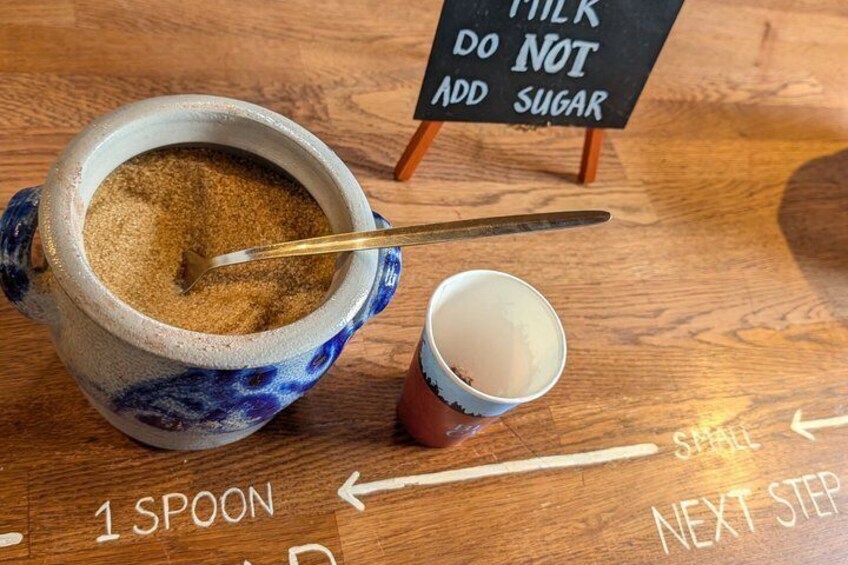 A ceramic jug with sugar, a spoon, and a cup of tea on a wooden table with a sign that says 'MILK DO NOT ADD SUGAR.' 4