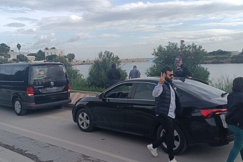 A black car parked on a road with a van in the background, near a body of water and greenery. 4