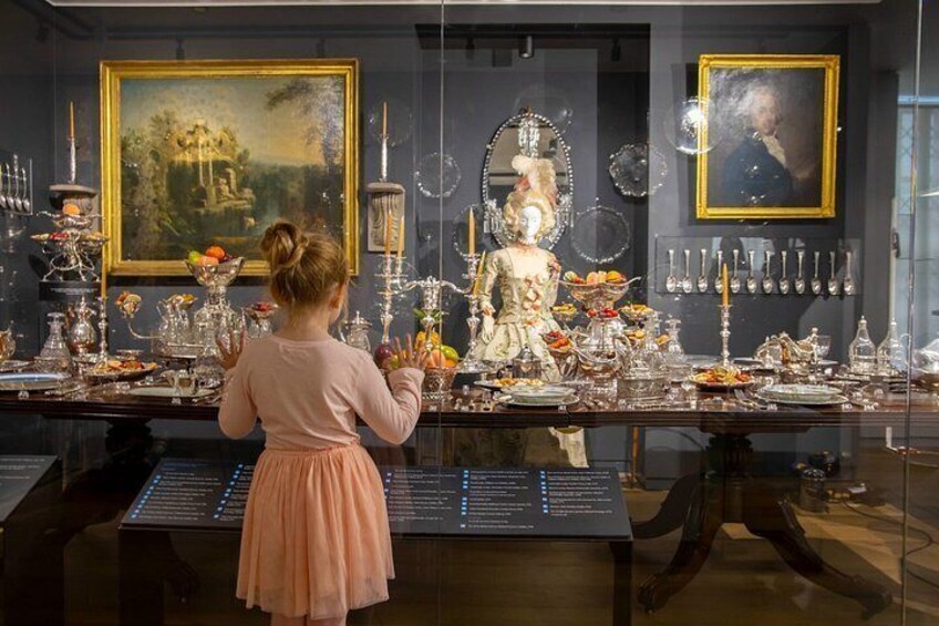 A child observing a display of antique silverware and glassware, with paintings and a dark-toned wall in the background. 2