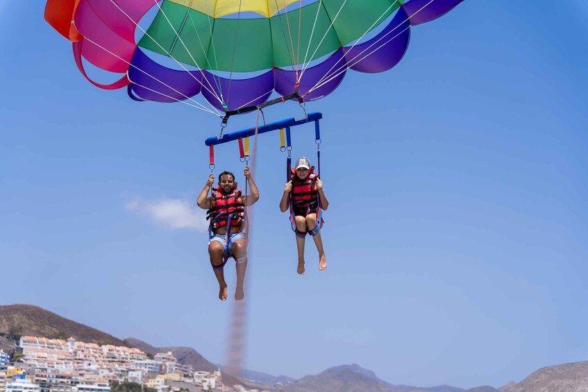 Two people skydiving with a parachute above them, with a coastal town and mountains in the background. 4