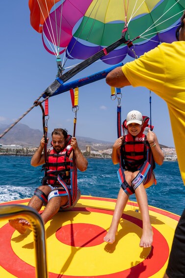 Two people parasailing over water with a colorful parachute, wearing life jackets and harnesses. 2