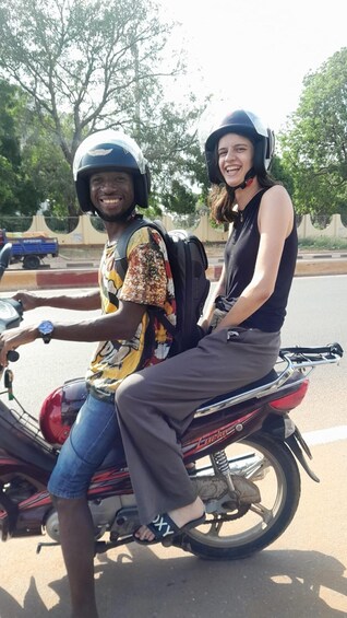 Two people on a motorcycle, one wearing a helmet, on a road with trees and a fence in the background. 2