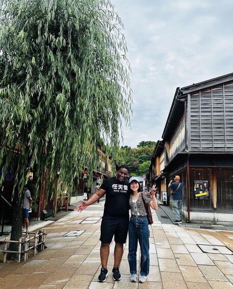 Two people standing in a pedestrian area with a weeping willow tree and traditional buildings. 3