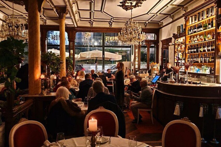 A busy restaurant interior with patrons dining, a bar area, and a view of the street outside. 1