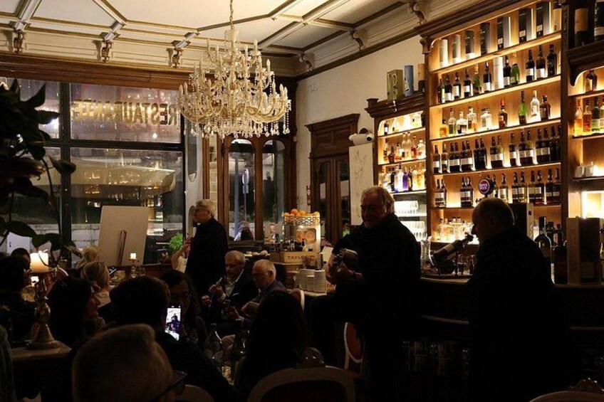 A bar with a chandelier, shelves of bottles, and people seated at tables. 4