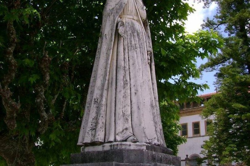 A weathered statue of a woman in a long dress, surrounded by trees and a building in the background. 1