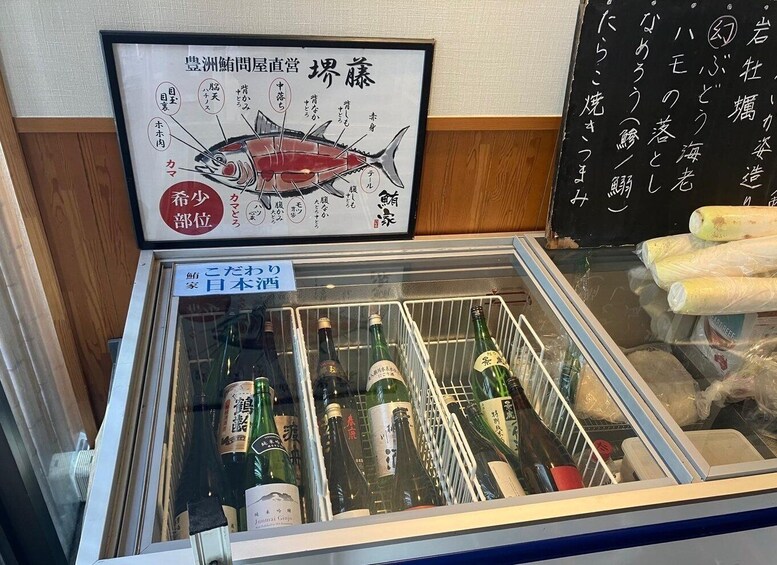 A refrigerated display case with bottles of Japanese liquor, a framed fish illustration, and a sign indicating the presence of a sushi bar. 2