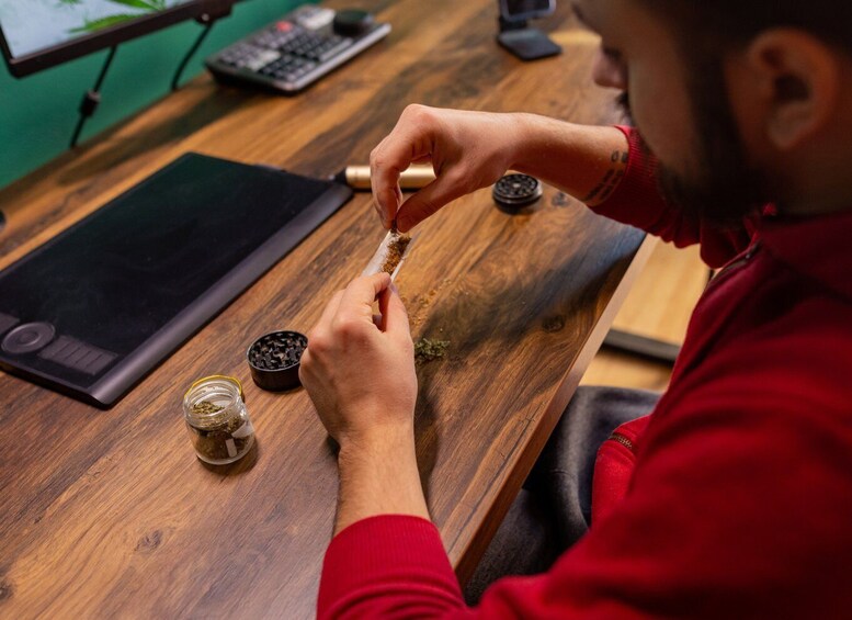 A person in a red shirt using a pipe, seated at a wooden table with a laptop, a calculator, and a bowl of herbs. 2