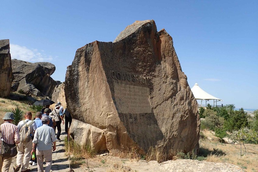 A large rock with inscriptions, a group of hikers, and a white pavilion in a dry landscape. 3