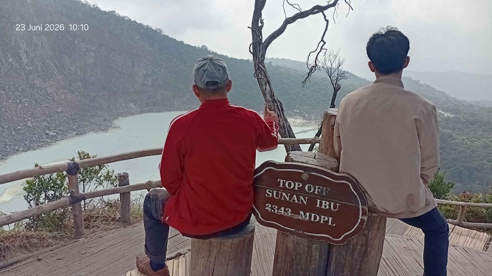 Two people sitting on a wooden bench overlooking a river, with a sign indicating the location as Top of Sunan Ibu MDPL. 1