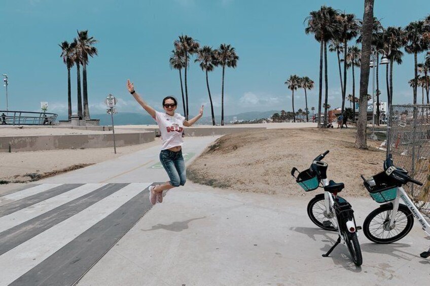 A person jumping in front of a bicycle on a beach with palm trees in the background. 1