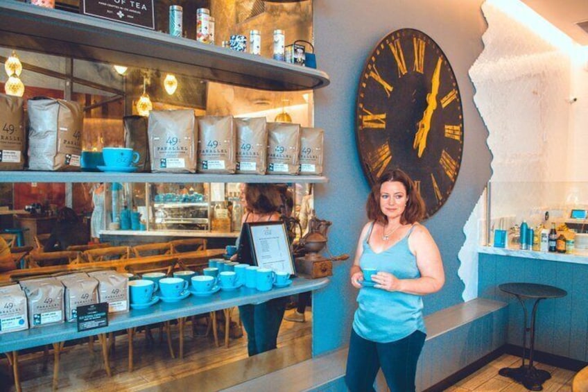 A person standing in a cafe with a large clock on the wall, coffee bags, and cups on shelves. 5