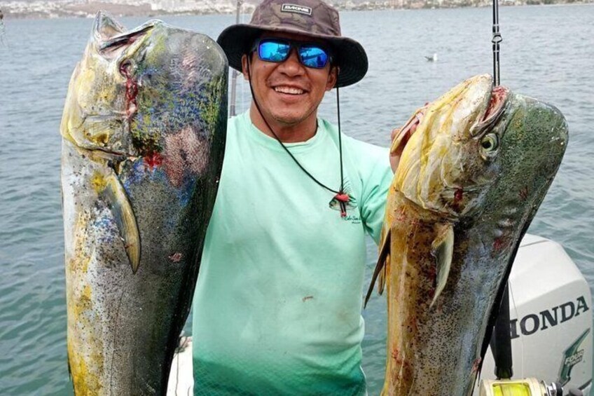 A person holding two large fish, one on each side, with a boat in the background. 1