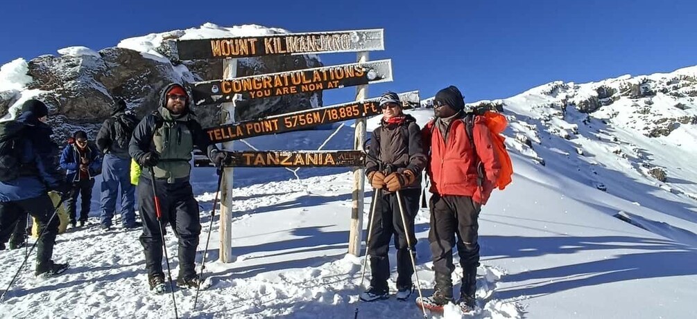 A group of people at a mountain summit with a sign indicating the elevation of 5756m/18,885ft and the location in Tanzania. 3