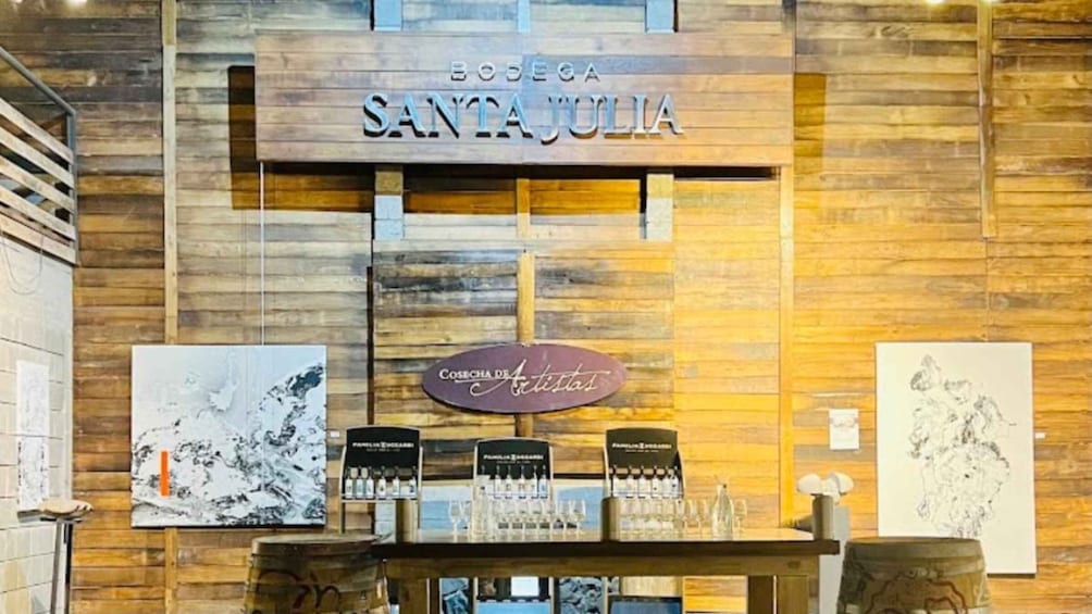Een houten wand met een bord waarop 'BODEGA SANTA JULIA' staat en een bord eronder met 'Cosecha de Artistas'. Er staan planken met glazen en een ingelijst kunstwerk op de wand. 2