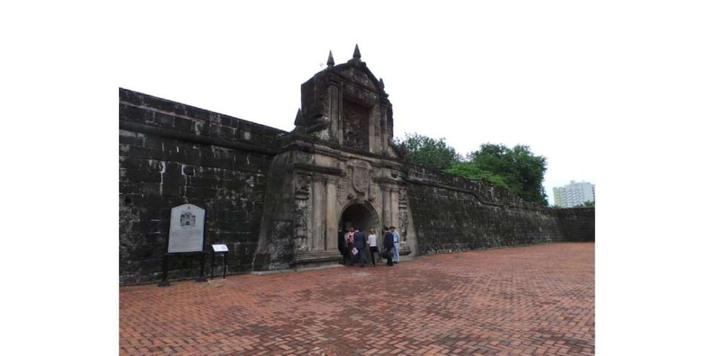 An ancient stone gate with a small tower, surrounded by a brick-paved area and a high wall. 1