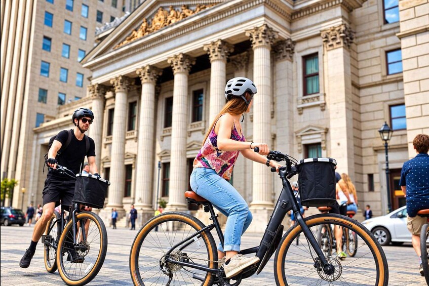Two people riding bicycles with baskets in front of a classical building with columns. 3
