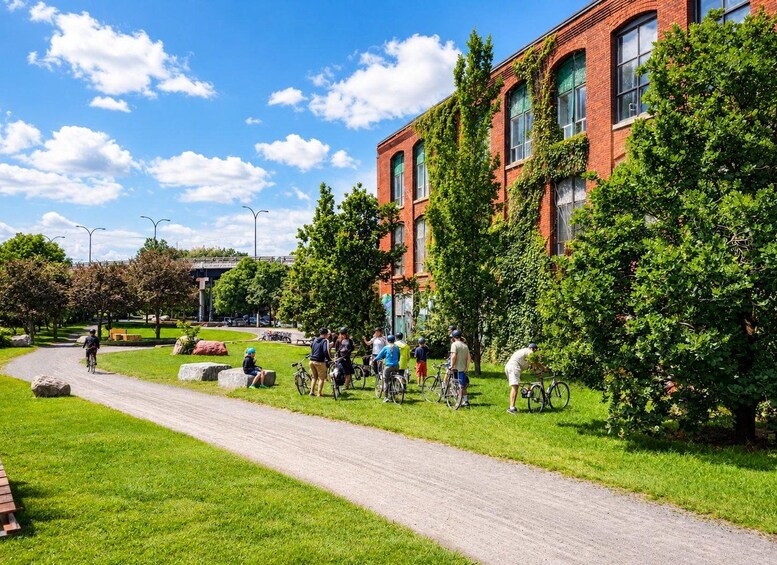 A group of cyclists on a paved path, a red brick building, and green trees. 2
