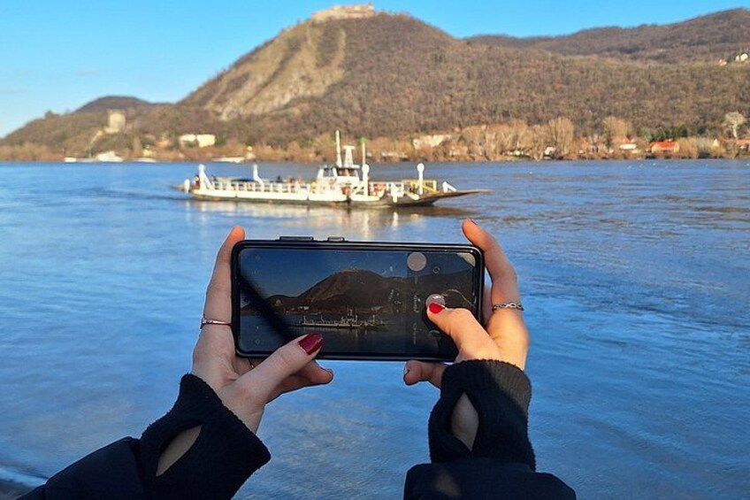 A person holding a smartphone, taking a photo of a boat on a body of water with mountains in the background. 1